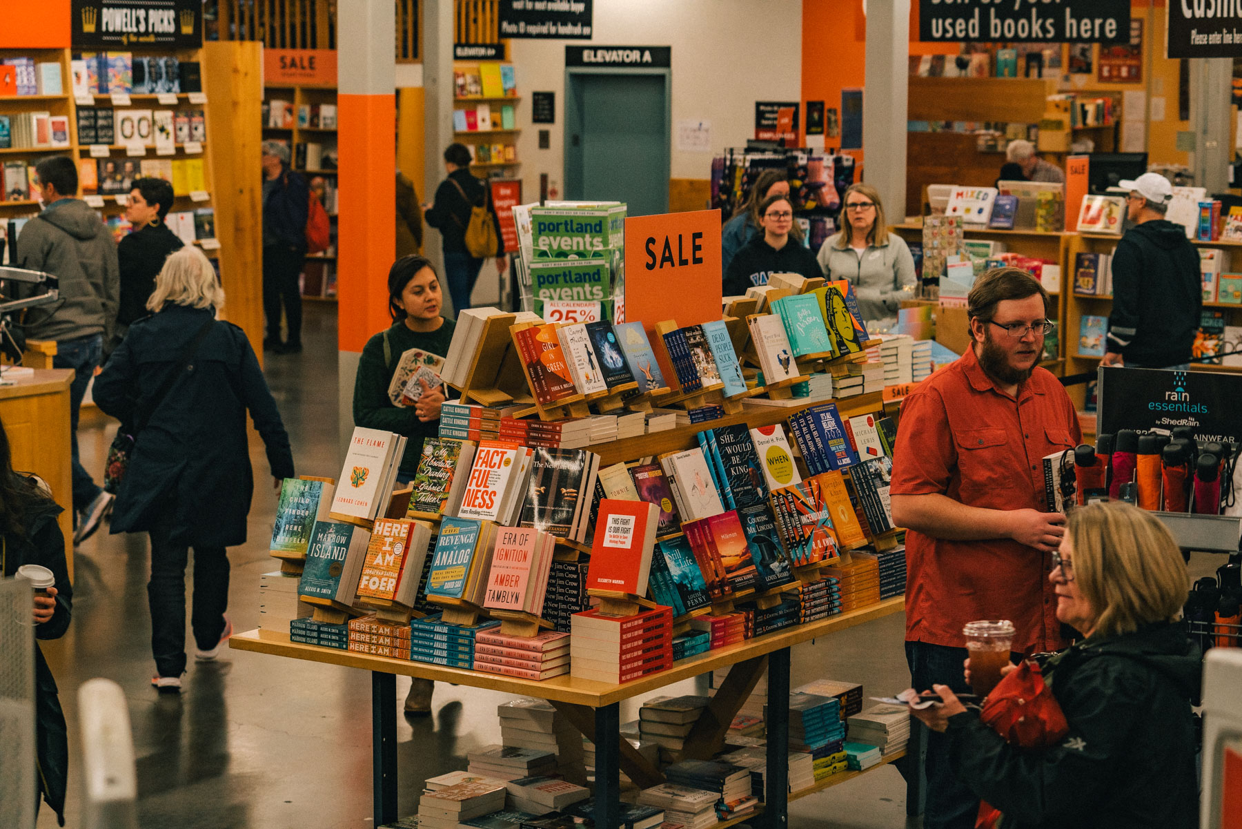 Shoppers in coats around a crowded sale table inside Powell's City of Books, with a rack of rain jackets and umbrellas on the right