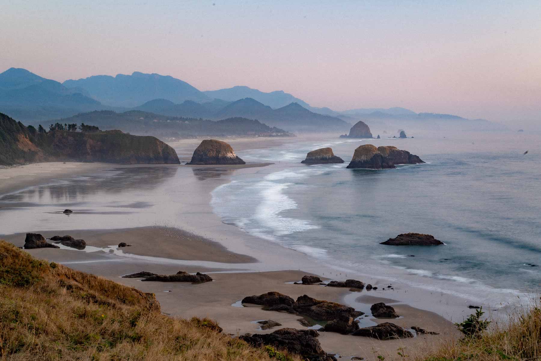 View south from the Ecola State Park headland in hazy pink light, with sea stacks standing in shallow surf, a long low-tide beach curving toward Haystack Rock and layered coastal mountains fading into the distance
