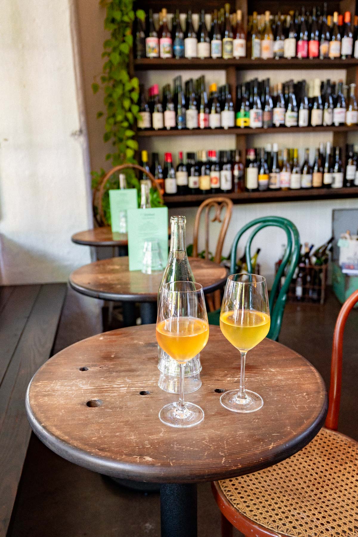 Two glasses of orange wine on a dark wood cafe table in front of a wall of wine bottles at a small Portland wine bar