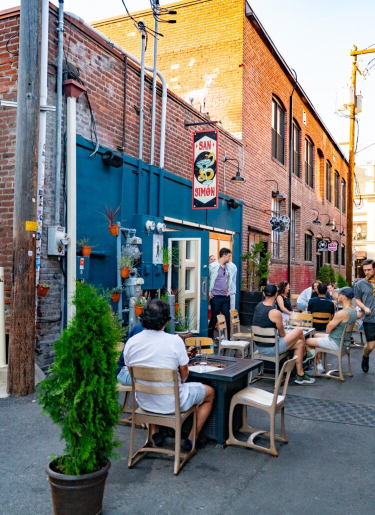 Diners at outdoor tables in an alley beside a brick building with a blue facade in Portland